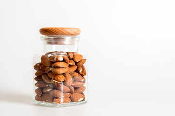 Close up. The glass jar is covered with a wooden lid and contains almonds inside. White background. Copy space.