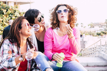 Playful real life concept with three cheerful smiling middle age women friends anejoying and laughing togehter playing with soap bubbles in outdoor leisure activity - playful and happiness concept