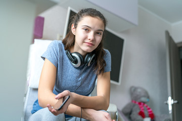 cute teen girl with headphones around her neck and phone in hand sits on a chair in the room and looks into the frame