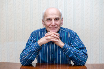 an older man sitting at a table with a smile, hands folded under face