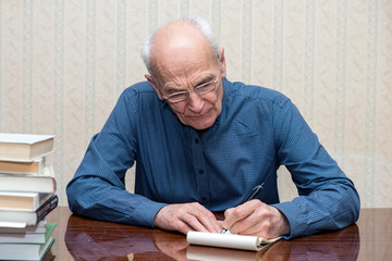 elderly man with glasses writes with his left hand in a notebook while sitting at the table, a stack of books near