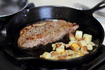 New York strip steak with potatoes frying in a cast iron pan on a natural gas stove top.