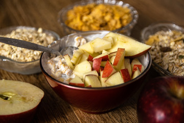Muesli with dried fruit, milk and sliced red apple on wooden table. Barley flake, fresh organic red apple,  and corn flakes in the background.