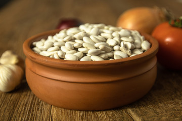 Uncooked white bean in a clay bowl on wooden table. Tomato, red onion, garlic, carrot and lettuce in the background.