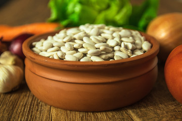 Uncooked white bean in a clay bowl on wooden table. Tomato, red onion, garlic, carrot and lettuce in the background.