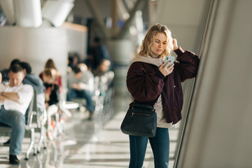 Curly blonde woman with phone in hands standing in waiting room at airport.