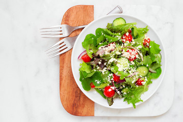 Healthy mixed salad with mixed greens, cucumber, onion, tomatoes and feta cheese. Top view on a marble and wood serving board.