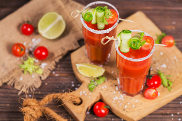 Two glasses of tomato juice decorated with fresh tomatoes, cucumber and leaves on a wooden background