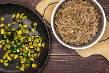 Mix boiled pasta with zucchini. recipe step by step fusilli pasta with zucchini on a frying pan with a stock pot on chopping board flatlay on brown wood