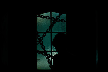 Dark silhouette of kid behind glass door with chain in supernatural green light. Locked alone in room behind door on chain on Halloween. Night kidnapping. Evil in home. Inside haunted house.