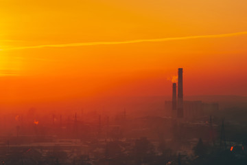 Fototapeta na wymiar Smog among silhouettes of buildings on sunrise. Smokestack in dawn sky. Environmental pollution on sunset. Harmful fumes from stack above city. Mist urban background with warm orange yellow sky.