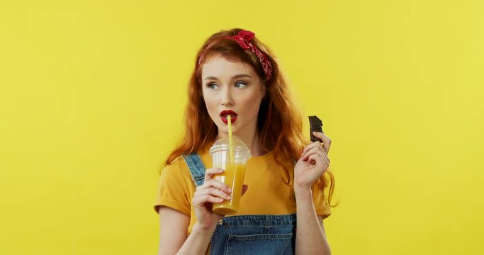 Caucasian young good looking and cheerful woman with red hair biting and eating chocolate cookie and drinking orange juice on the yellow background.