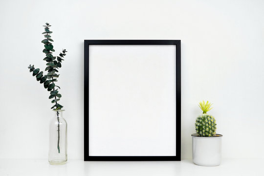 Mock up black frame with cactus and branches on a shelf or desk. White shelf and wall. Portrait frame orientation.