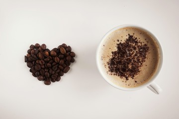  white cup with cappuccino and frothy and grated chocolate on a beige background and a heart of coffee beans to the left of the cup
