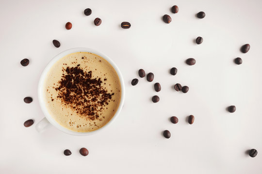 white cup with cappuccino and crema and grated chocolate on a beige background and roasted coffee beans around the cup