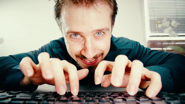 Man typing on keyboard with evil laugh stare into camera 4K. Low angle view from behind the keyboard with a male person face in focus while typing.