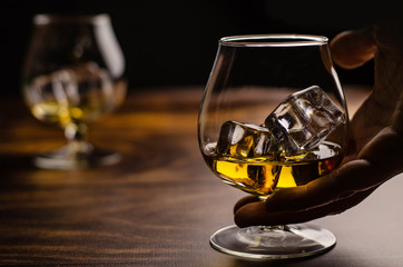 Whiskey / cognac glass with ice in a hand on a wooden background. Dark backdrop.