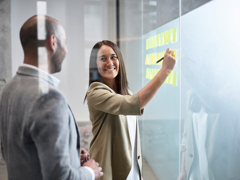 Businesswoman smiling at colleague writing on sticky notes at glass pane in office - Powered by Adobe