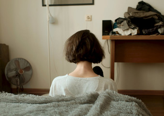 Brunette girl with a short cut, sitting with her back to the camera in a messy room. Loneliness,...
