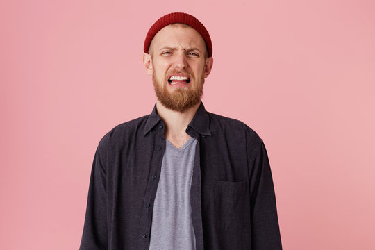 Ugh, how disgusting! Displeased young bearded male, with red hat, says fie and has dissatisfied facial expression as sees something abominable or detestable, looking at camera over pink background.