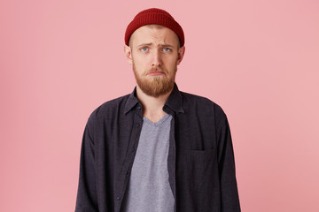 Close up of a sad bearded man about to cry, in blak shirt and t-shirt, with red hat, looking at the camera and frowning over pink background.