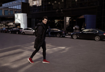 Young man crossing the road of city street. Wearing in black jacket and trousers, red sneakers, backpack, headphones. Fashion style