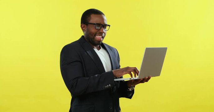 Attractive African American young businessman in glasses standing on the yellow background with a laptop computer and tapping or typing.