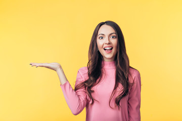 Beautiful smiling woman holding and presenting copy space on her palm isolated on yellow background