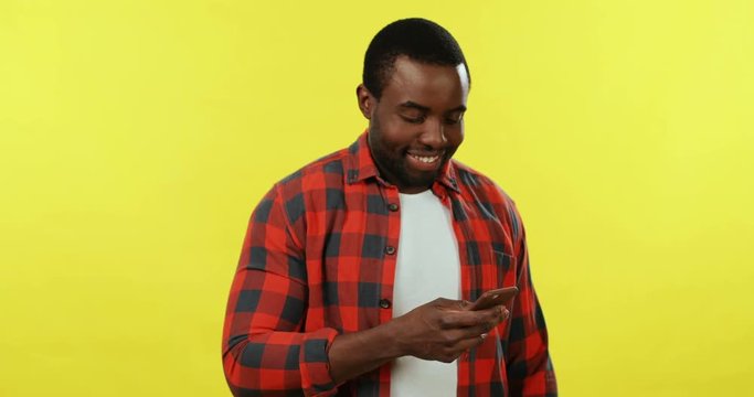 Young African American handsome guy standing on the yellow background and tapping or scrolling on the smartphone.