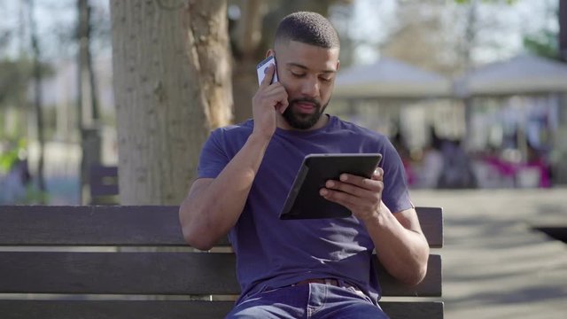 Young Afro-American short-cut muscular man with stylish beard in blue T-shirt sitting on bench in park, holding tablet in hands, talking on phone. Front view. Communication, lifestyle concept