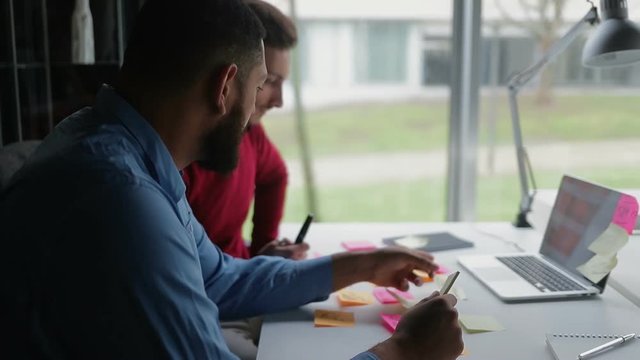 Two young men sitting at table with sticky notes and laptop. Confident businessmen working at office. Business and technology concept