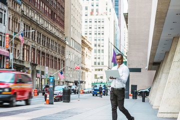 Obraz premium Young African American businessman traveling, working in New York, wearing white shirt, black pants, holding laptop computer, walking on street, looking down, reading. Cars, buildings on background.