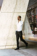 Young African American businessman traveling, working in New York, wearing white shirt, black pants, leather shoes, carrying laptop computer, walking out from office building, talking on cell phone..