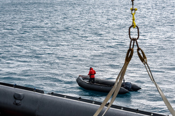 Black inflatable boat in water with man in red coat, second boat being winched off ship, Atlantic Ocean