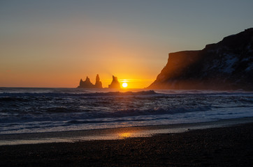 Sunset behind jagged cliffs, over the ocean waves, and black sand beach of Vik Iceland