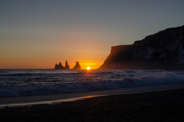 Sunset behind jagged cliffs, over the ocean waves, and black sand beach of Vik Iceland