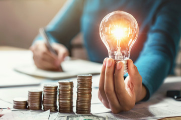 business woman hand holding lightbulb with coins stack on desk. concept saving energy and money