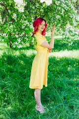 Beautiful and young woman with bright red hair and ref lips standing next to a blooming apple tree in a yellow dress.