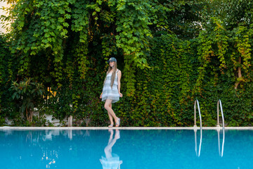 A young girl stands on the edge of the pool in a white dress on a background of green plants