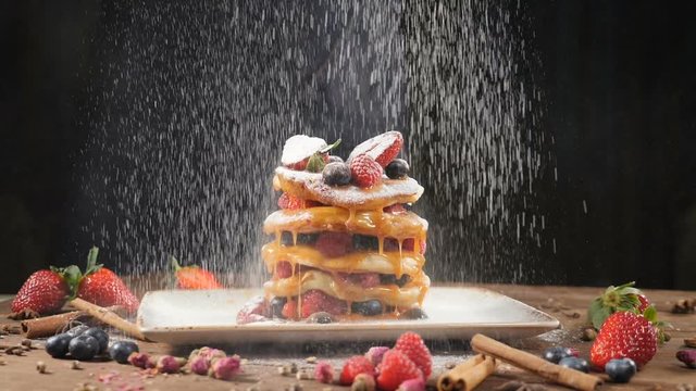 Chef Sprinkling sugar powder on tasty pancake, slow motion. Flour being sieved onto pancakes. Close-up pancakes stack on plate being decorated with cranberries and dusted with powdered sugar