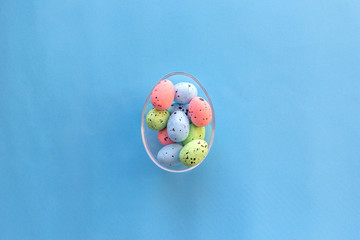 Colorful quail eggs in large glass egg on blue background. Happy Easter concept. Top view, flat lay.