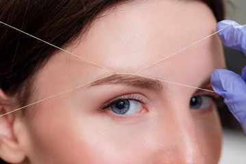 Cosmetologist plucks client eyebrows by thread. Close up
