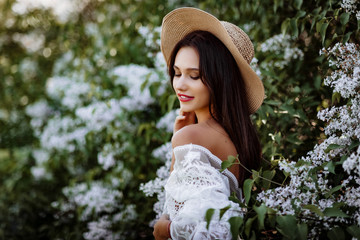 young woman in a straw hat in lilac. Model in white pennuar among flowers