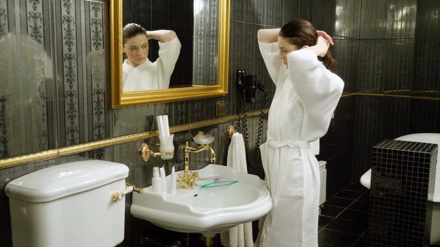 Young beautiful woman dressed in bathrobe looking at her reflection in bathroom mirror. 4K