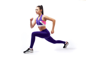 A dark-haired woman coach in a sporty purple  short top and gym leggings makes lunges  by the feet forward, hands are held out to the side   on a  white isolated background in studio