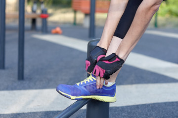 Female in sportswear and protective gloves ties bright sneakers preparing for a jog, run or other fitness. Morning workout on outdoors background. Copy space.