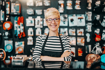 Portrait of beautiful smiling Caucasian female worker with short blonde hair standing in bicycle shop with arms crossed. - Powered by Adobe