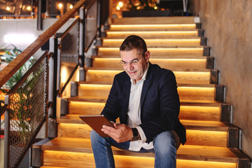 Middle aged man dressed smart casual sitting on the stairs in cafeteria and using tablet.