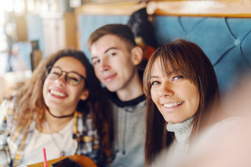 Three multicultural teenage classmates taking selfie while sitting at cafeteria.