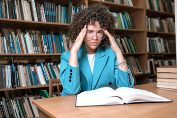 Young beautiful curly girl in glasses and blue suit sitting with books in the library. Student Study